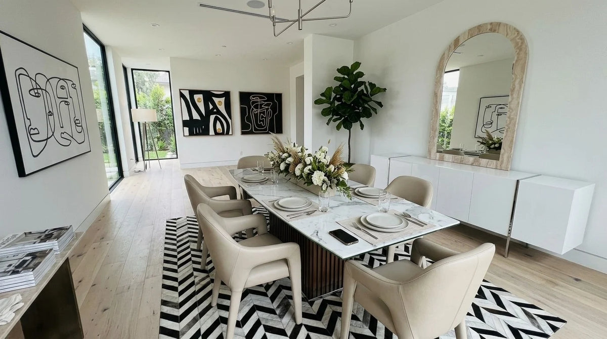 Modern dining room with a white marble-look table on a fluted walnut pedestal, cream leather armchairs, a black and white herringbone cowhide rug, line art and a white buffet