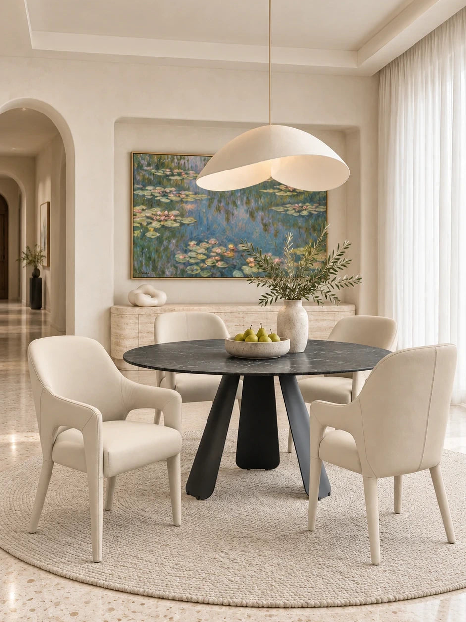 Round black ceramic dining table on a matte black blade base with four cream leather dining armchairs and a travertine buffet in a bright dining room