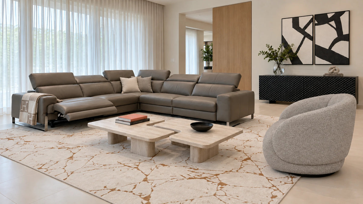 Living room with a taupe leather reclining sectional, a two-piece travertine coffee table, an ivory rug with copper veining, a gray swivel chair, black and white art and a textured black buffet